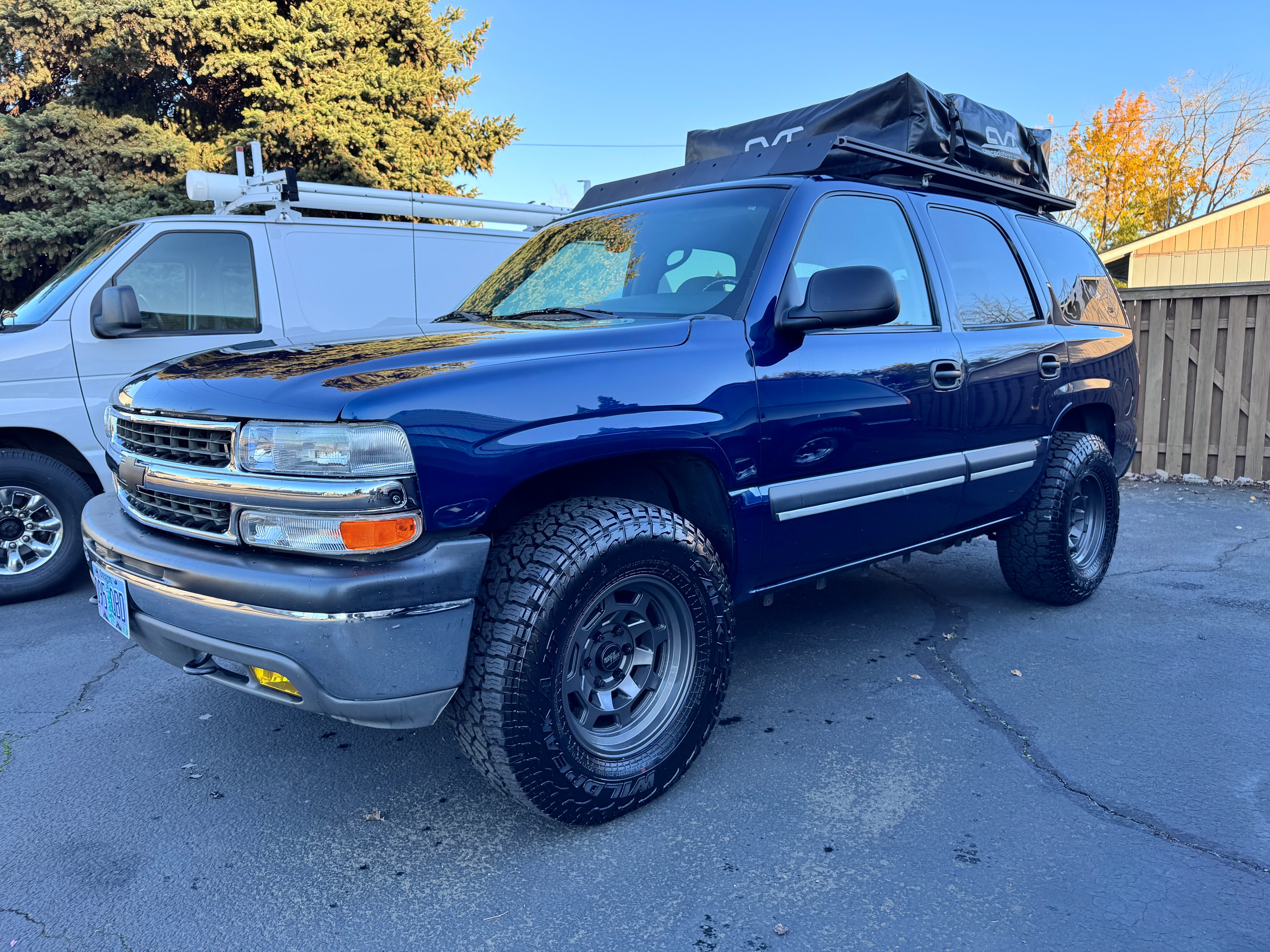 Front 3/4 view of a dark blue 2003 Chevrolet Tahoe LS in golden-hour light with a custom aluminum roof platform and CVT rooftop tent, on gunmetal wheels and 33-inch Falken Wildpeak all-terrain tires