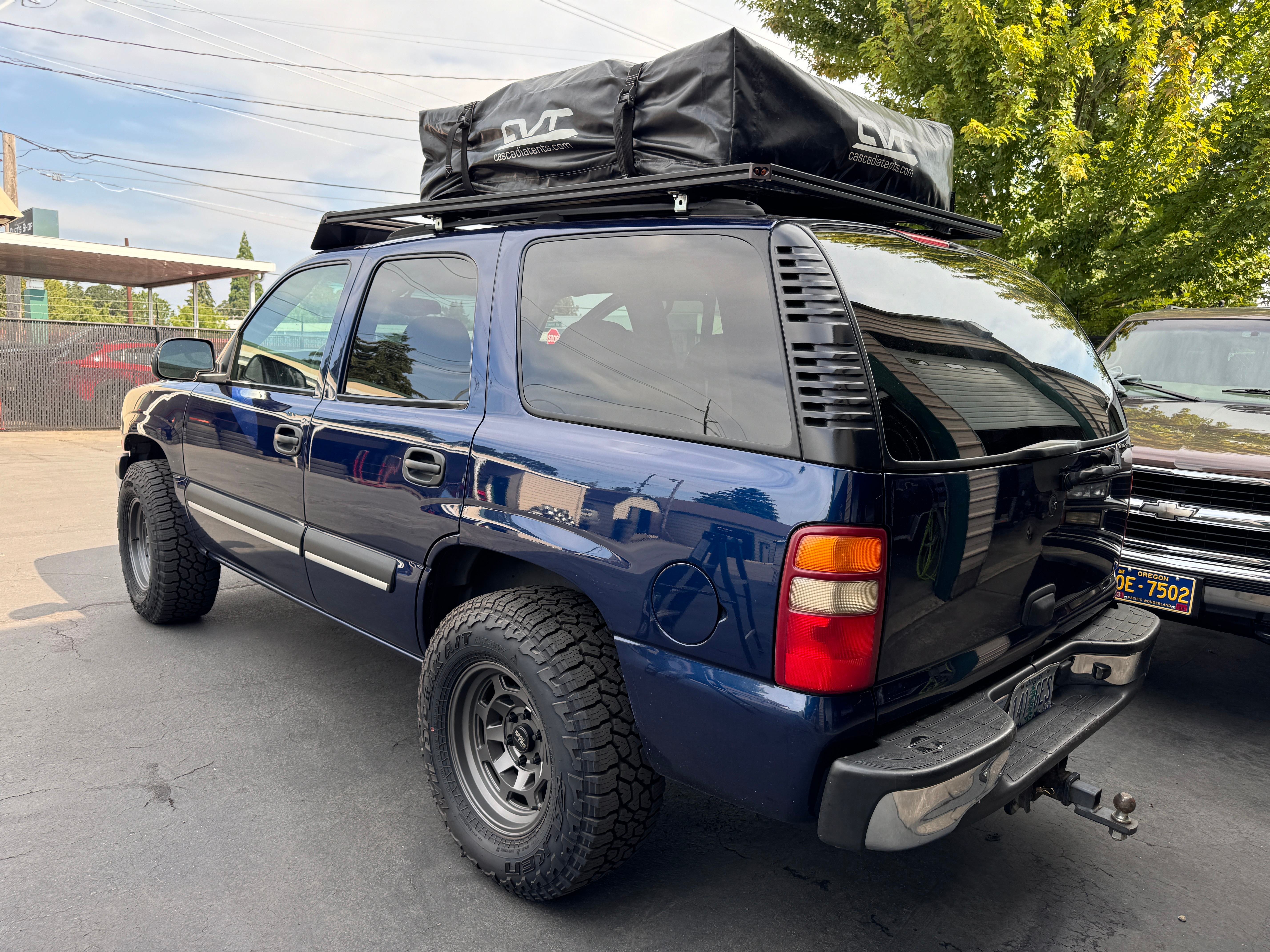 Dark blue 2003 Tahoe LS with a rooftop tent folded on a custom aluminum roof platform, parked outside on gunmetal wheels and Falken all-terrains