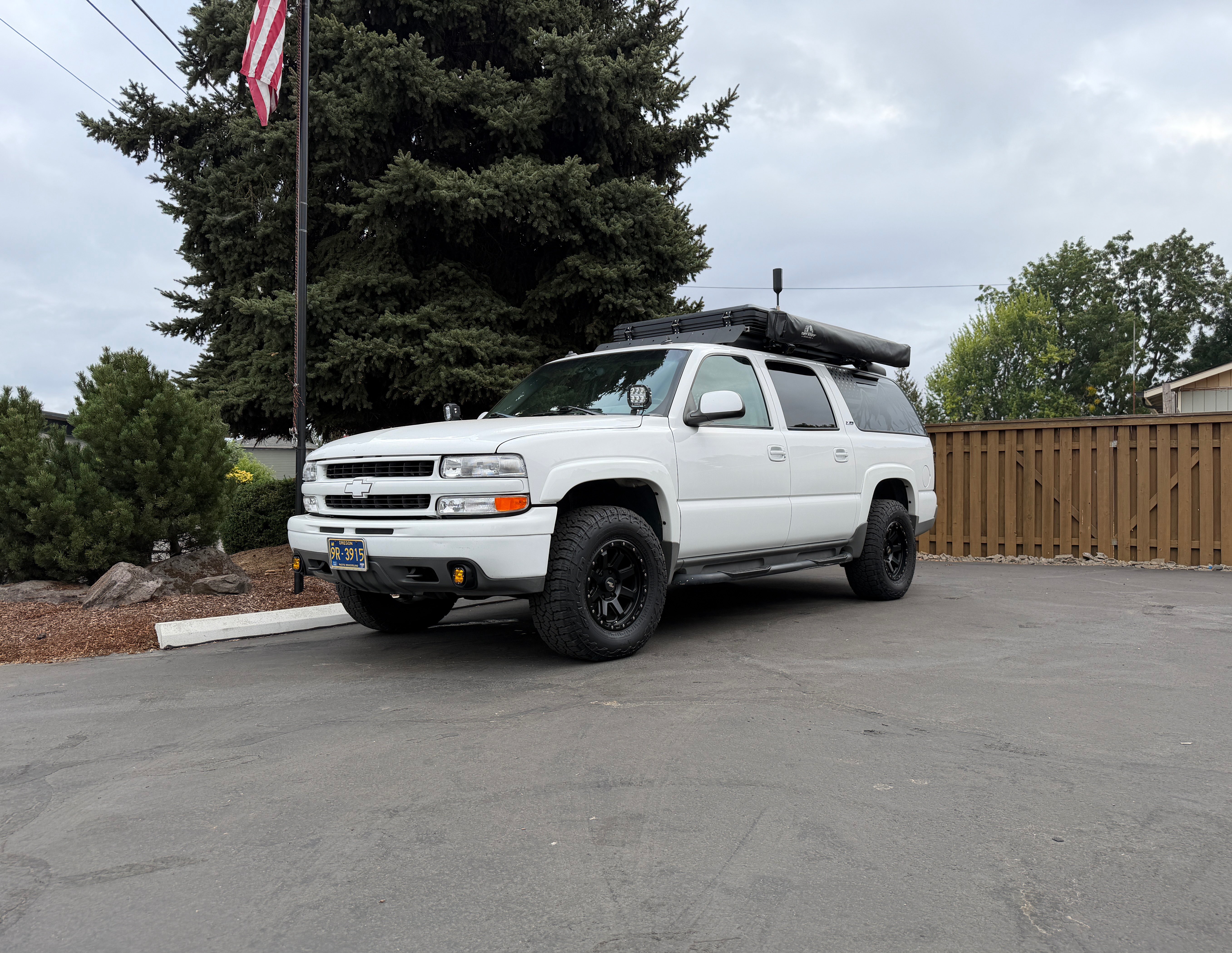 White 2003 Chevrolet Suburban Z71 with a roof rack, rooftop tent, and awning on black wheels and 32-inch all-terrain tires