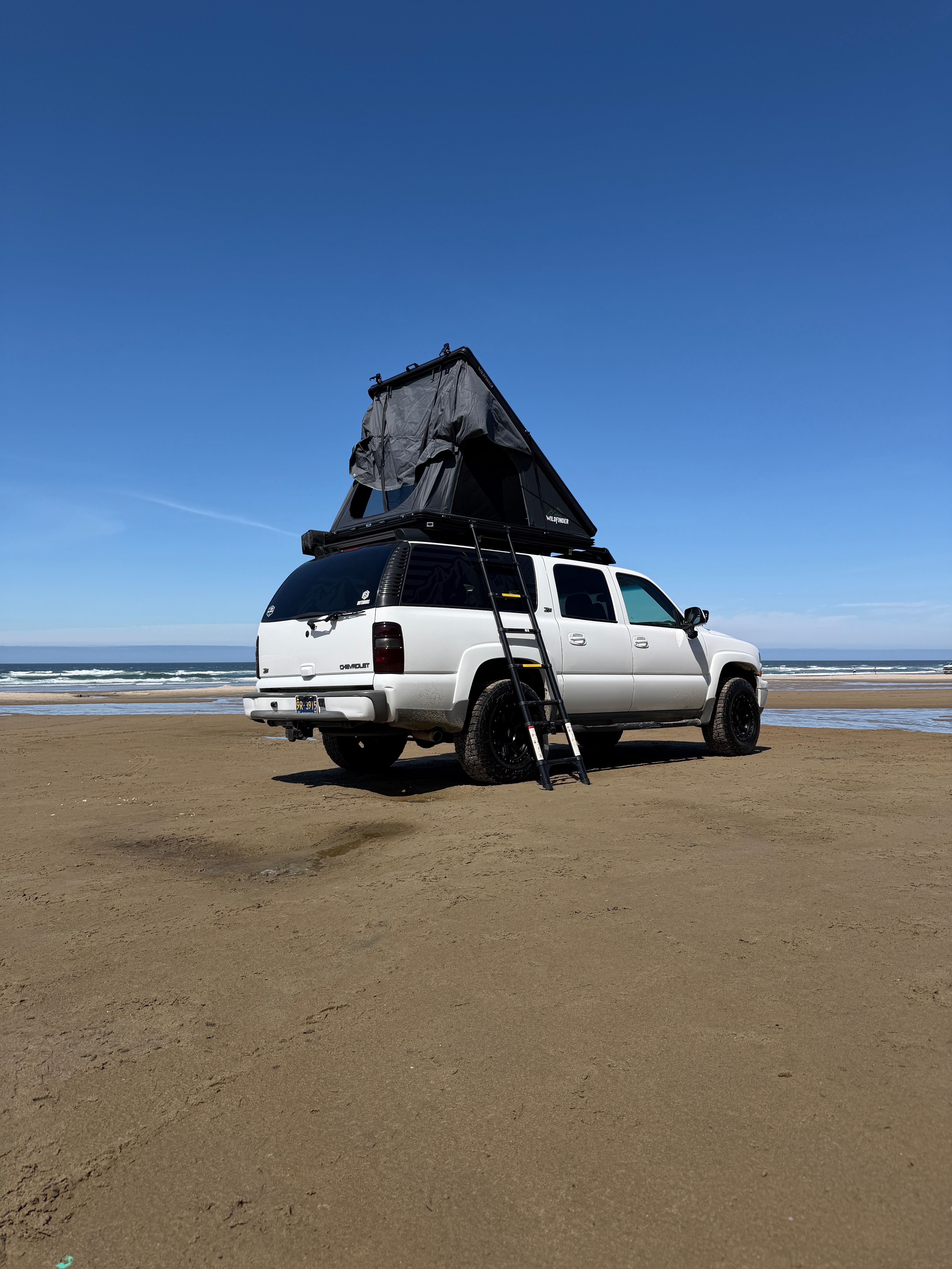 White Suburban Z71 on the Oregon coast with a WildFinder hard-shell rooftop tent popped open on a custom roof platform, ladder extended to the sand